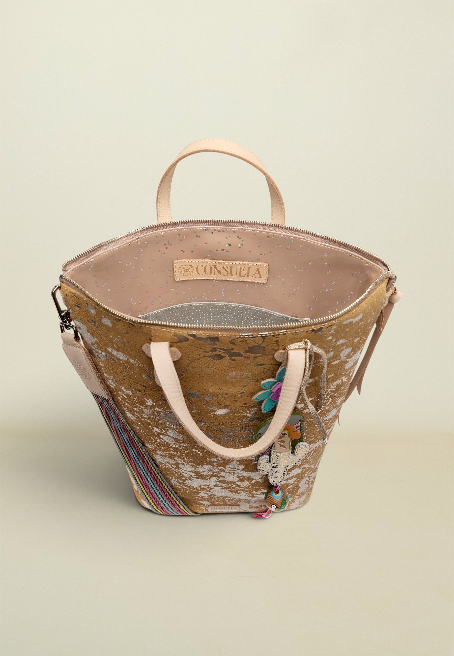 A Consuela handbag with a distressed brown and beige pattern, featuring light pink handles, a striped shoulder strap, and colorful embroidered details, set against a solid light beige background.