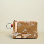 A small zip pouch with a brown and beige abstract paint splatter pattern, featuring a light pink leather zipper pull, a silver metal key ring, and a small rectangular brand patch on the front.