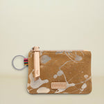 A small zippered pouch with a brown suede-like finish and a white and gray paint splatter pattern. It features a light tan leather zipper pull with the brand name 'CONSIDAS' and a colorful braided key ring attached on the left, set against a solid light beige background.