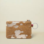 A small zippered pouch with a tan and white cowhide pattern, featuring a silver zipper, a small colorful striped pull tab, and a clear plastic key ring, set against a solid light beige background.