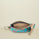 A small zippered pouch with a colorful abstract pattern in turquoise, pink, orange, and green, featuring a beige leather zipper pull and a silver key ring, shown open on a light beige background.