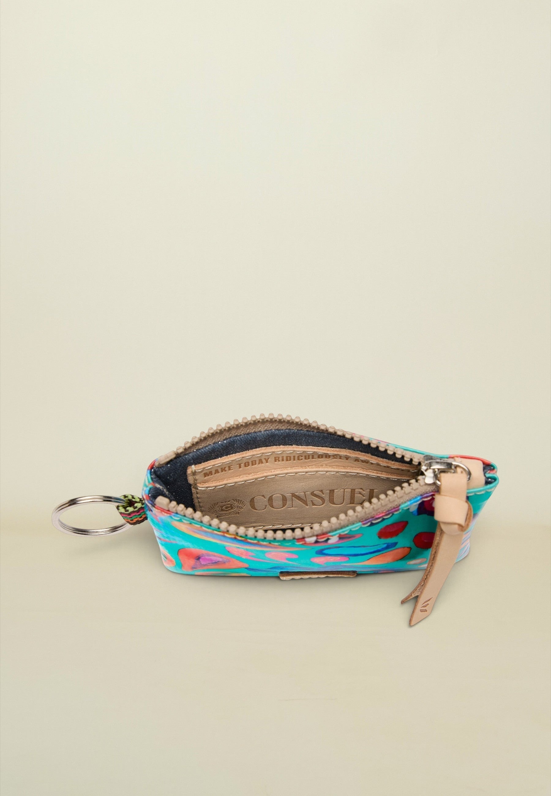 A small zippered pouch with a colorful abstract pattern in turquoise, pink, orange, and green, featuring a beige leather zipper pull and a silver key ring, shown open on a light beige background.