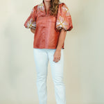 A woman modeling a rust-colored short-sleeve blouse with voluminous patterned puff sleeves and white pants, standing against a plain beige studio background.