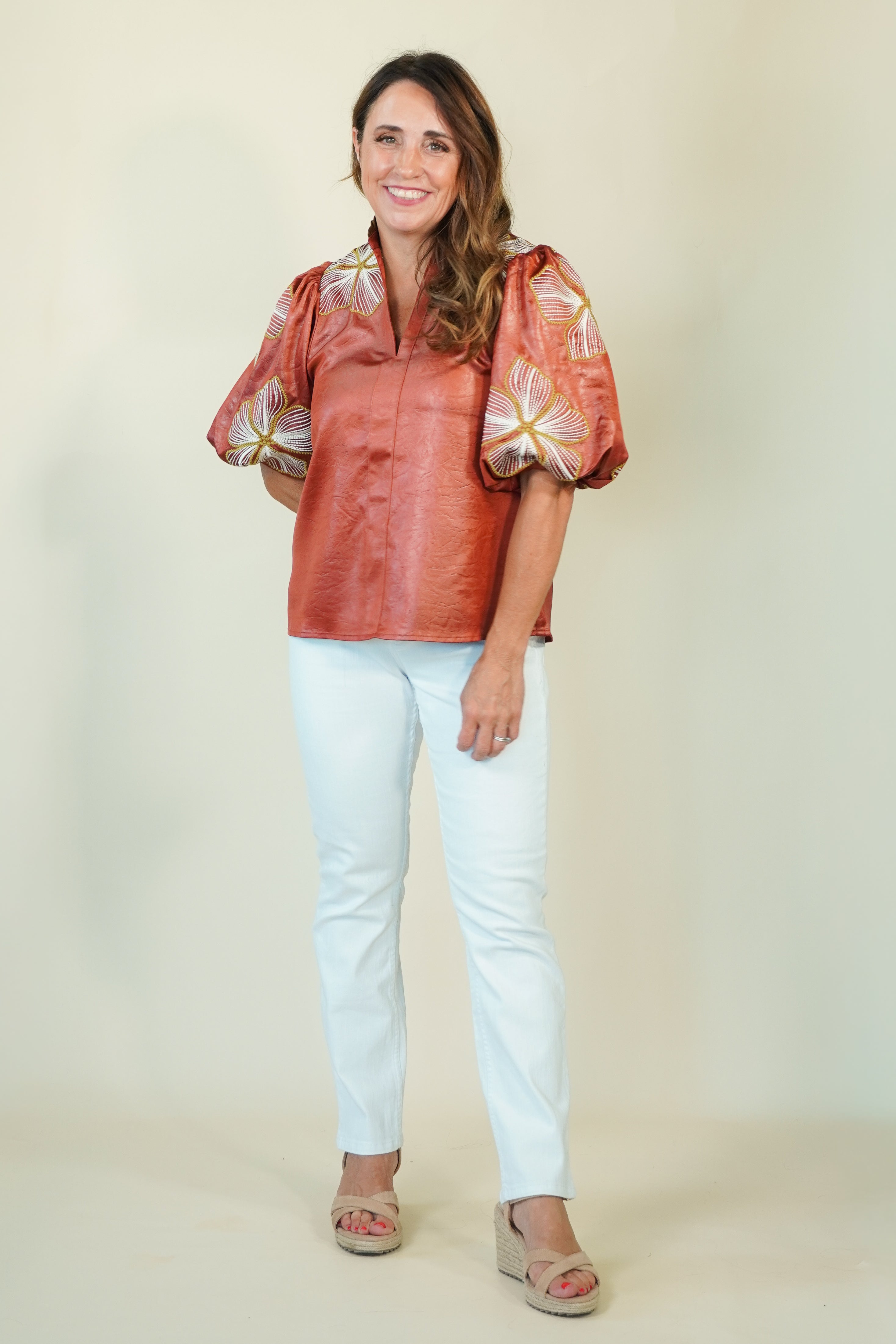 A woman modeling a rust-colored short-sleeve blouse with voluminous patterned puff sleeves and white pants, standing against a plain beige studio background.