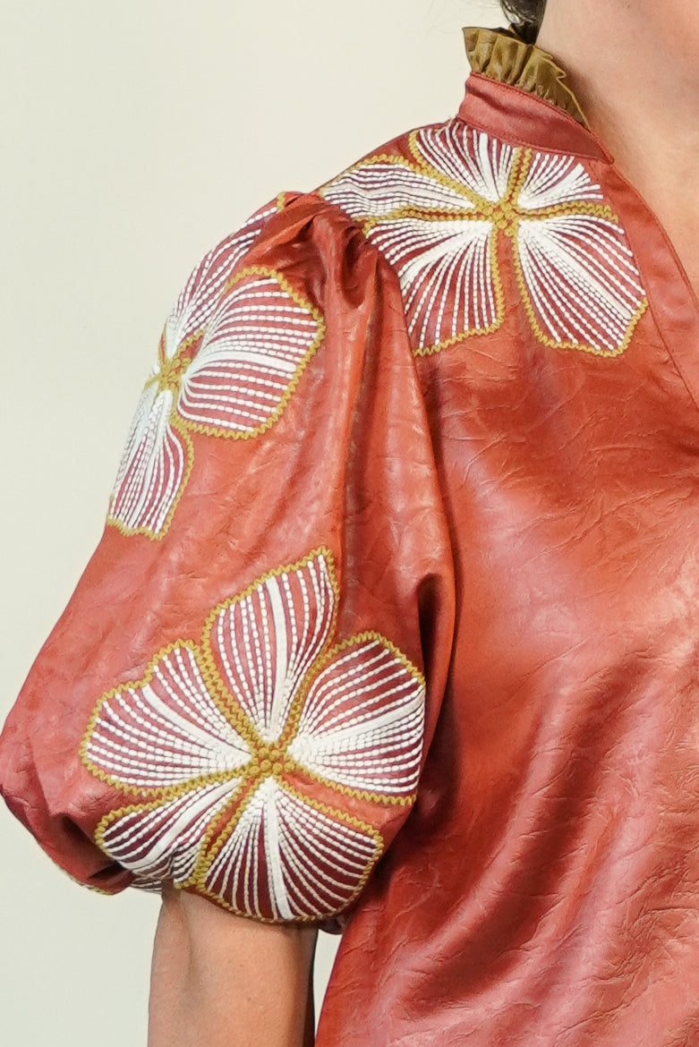 Close-up of a terracotta-colored satin blouse with puff sleeves, featuring large white and gold embroidered floral motifs on the shoulders and upper chest.