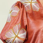 Close-up of a terracotta-colored satin blouse with puff sleeves, featuring large white and gold embroidered floral motifs on the shoulders and upper chest.