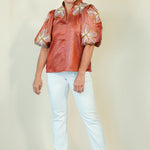 A woman modeling a short-sleeved metallic copper-brown blouse with large white and gold floral puff sleeves, paired with light blue jeans and beige wedge sandals, against a plain cream background.