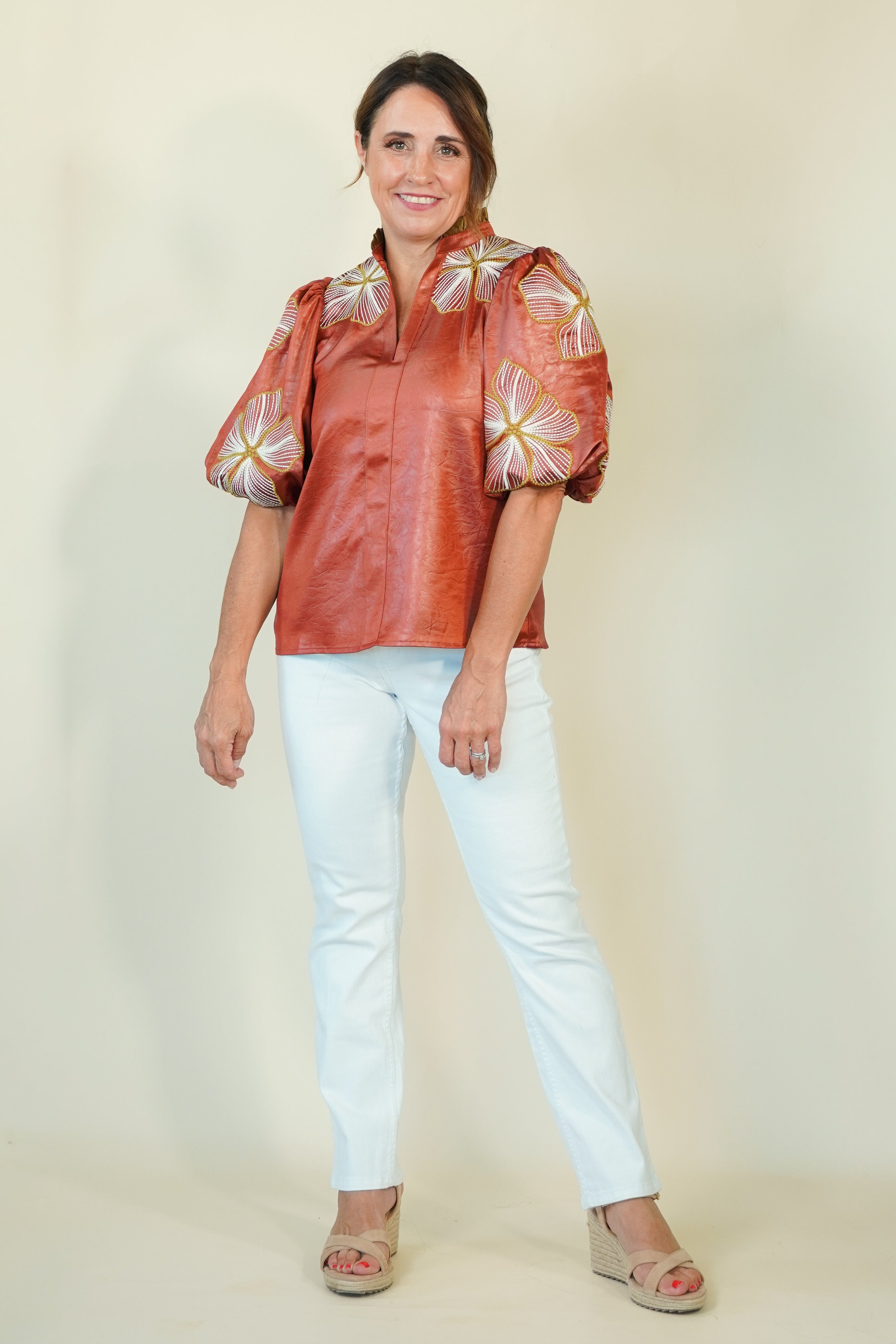 A woman modeling a short-sleeved metallic copper-brown blouse with large white and gold floral puff sleeves, paired with light blue jeans and beige wedge sandals, against a plain cream background.