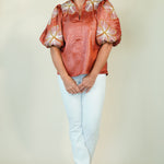 A woman modeling a rust-orange satin blouse with white and gold floral puff sleeves, paired with light blue jeans and beige sandals, against a plain beige studio background.