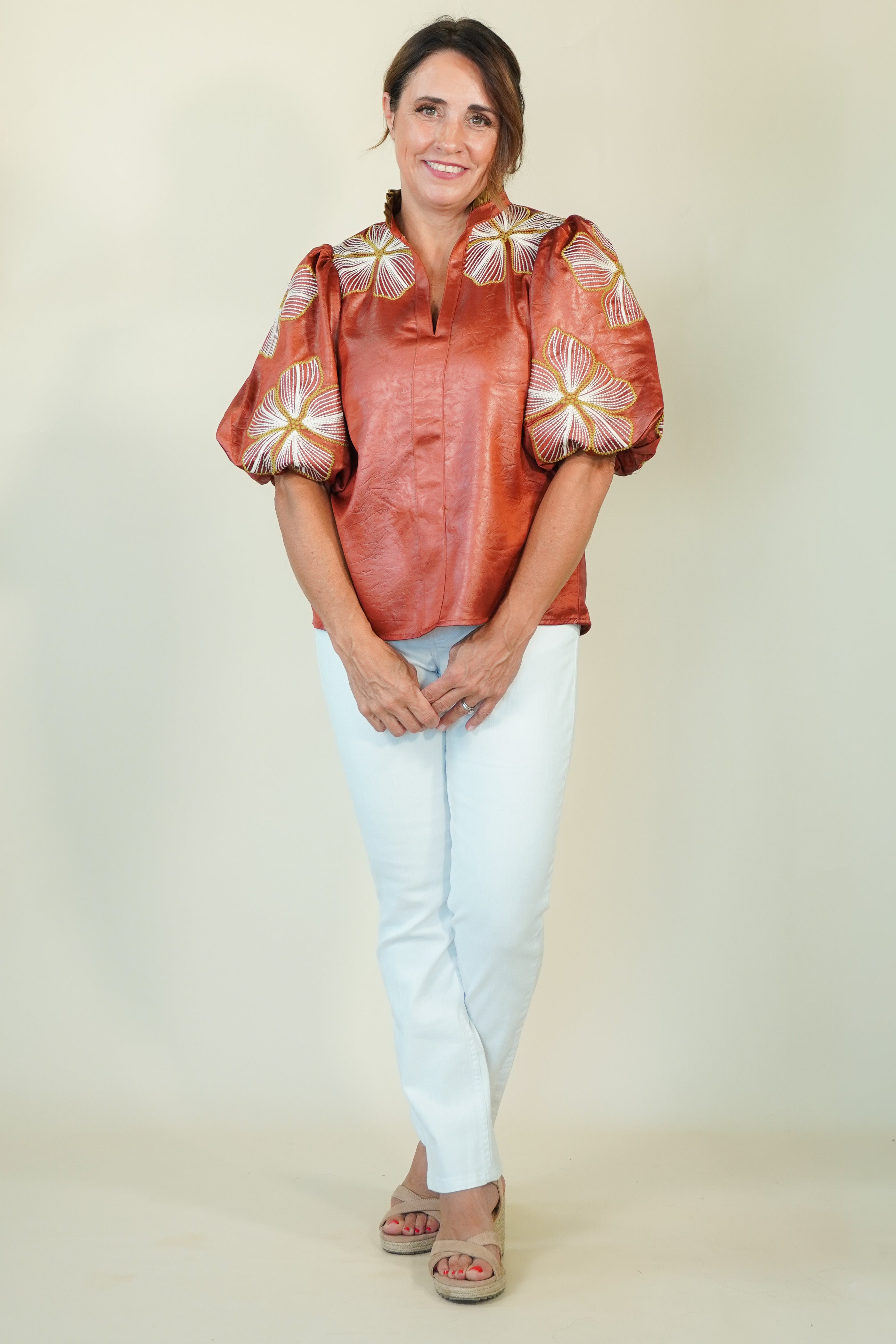 A woman modeling a rust-orange satin blouse with white and gold floral puff sleeves, paired with light blue jeans and beige sandals, against a plain beige studio background.