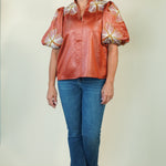 A woman modeling a short-sleeve button-up blouse with puffed sleeves in a rust-orange color, decorated with white and yellow floral patterns on the shoulders and sleeves. She is wearing blue jeans and beige sandals, standing against a plain light beige studio background.