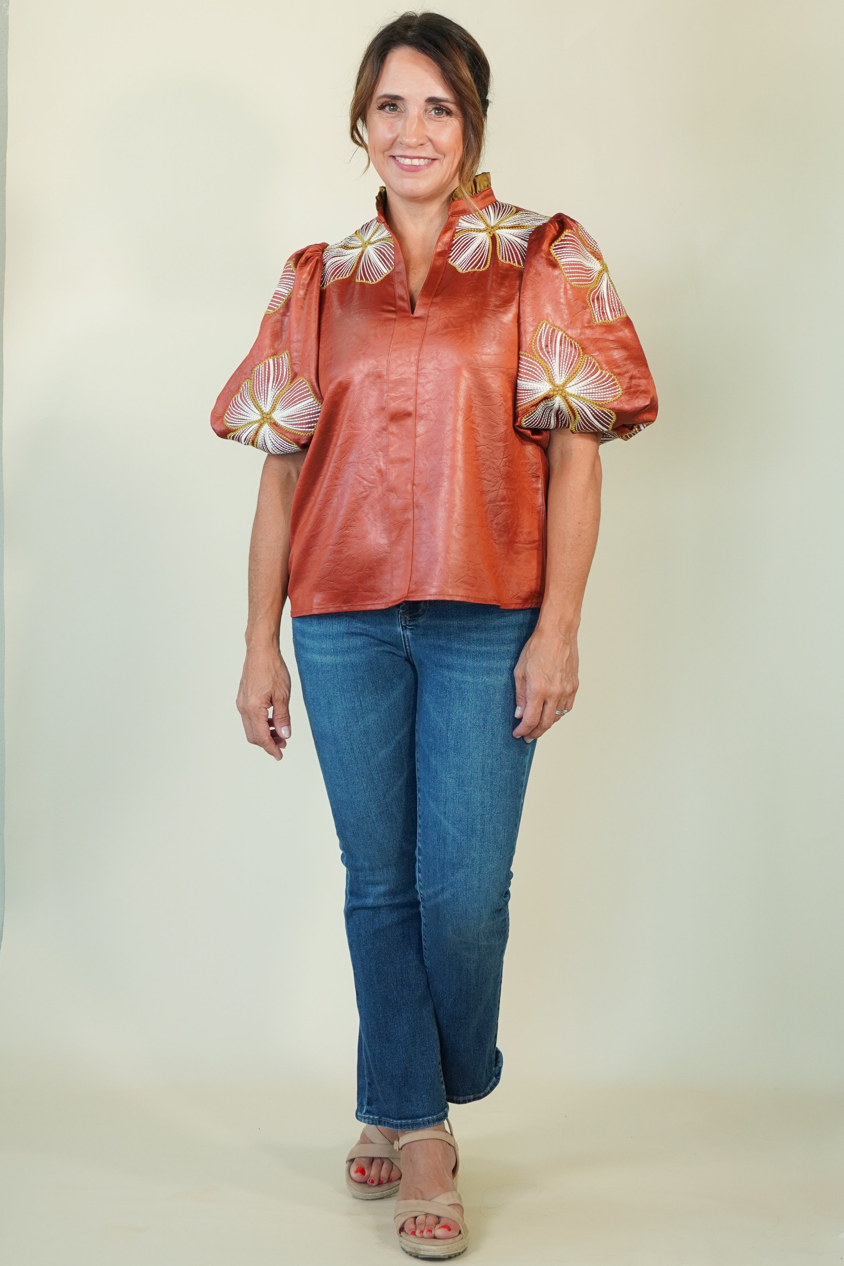A woman modeling a short-sleeve button-up blouse with puffed sleeves in a rust-orange color, decorated with white and yellow floral patterns on the shoulders and sleeves. She is wearing blue jeans and beige sandals, standing against a plain light beige studio background.