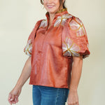 A woman modeling a short-sleeve collared blouse with a glossy rust-orange finish and large white floral motifs with gold centers on the shoulders and sleeves, paired with blue jeans against a plain light beige studio background.