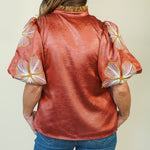 A woman viewed from the back wearing a metallic copper-orange blouse with puffed sleeves featuring white and gold floral embroidery, paired with blue jeans against a plain light beige background.
