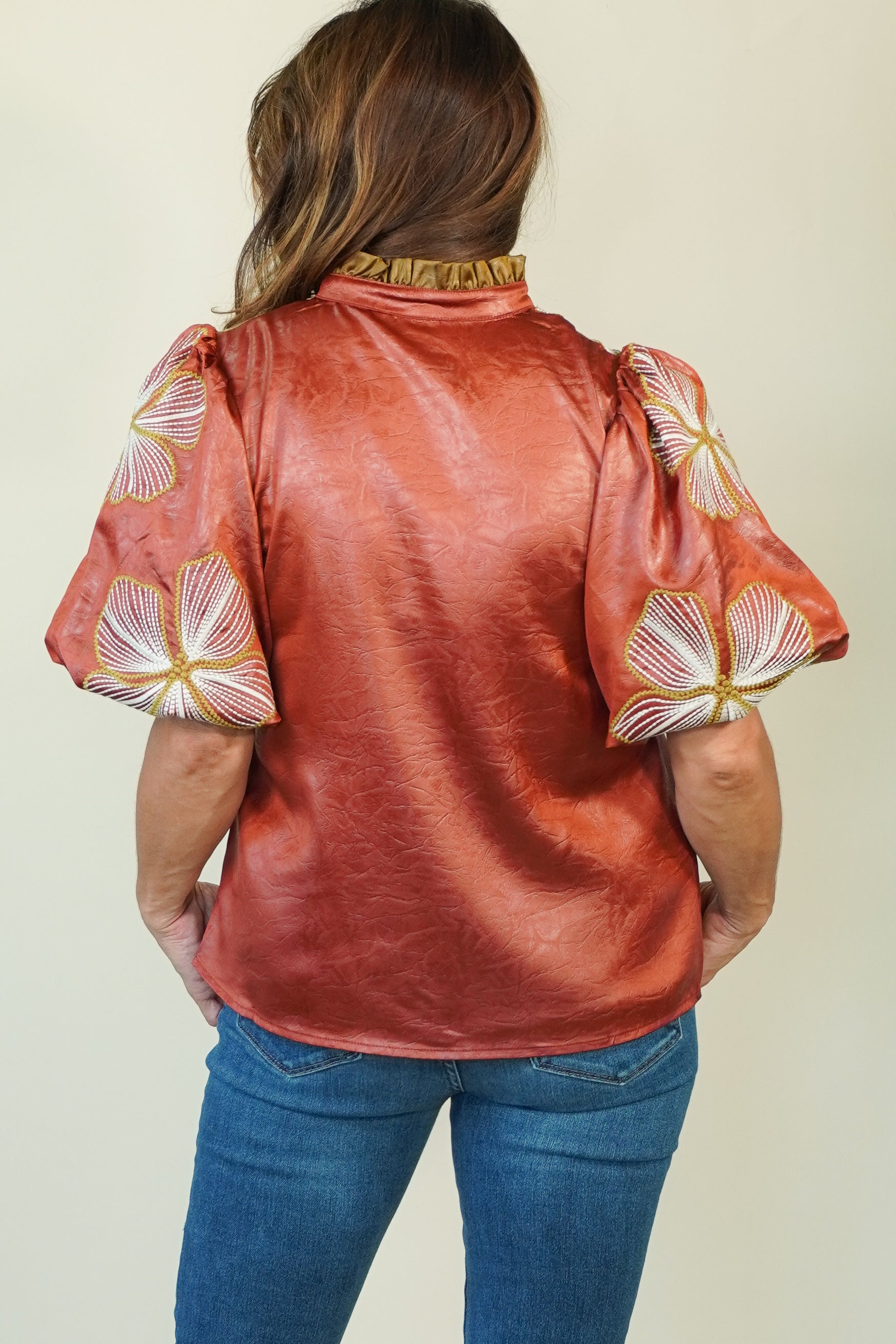 A woman viewed from the back wearing a metallic copper-orange blouse with puffed sleeves featuring white and gold floral embroidery, paired with blue jeans against a plain light beige background.