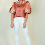 A woman modeling a copper-orange satin blouse with puffed sleeves and white floral embroidery, paired with light blue jeans and beige wedge sandals, against a plain cream background.