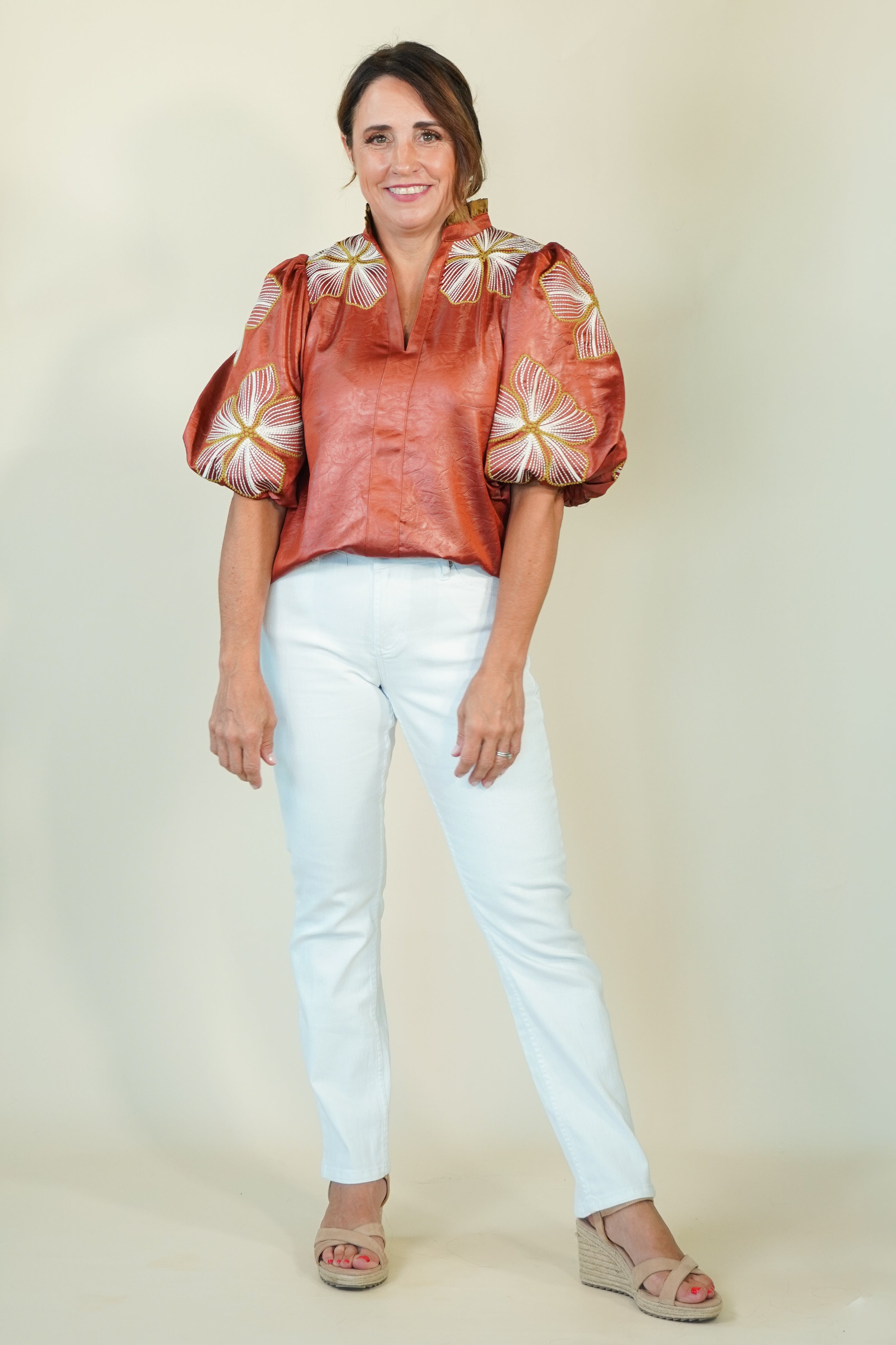 A woman modeling a copper-orange satin blouse with puffed sleeves and white floral embroidery, paired with light blue jeans and beige wedge sandals, against a plain cream background.