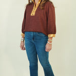 A woman modeling a rust-brown long-sleeve top with gold patterned trim on the collar and cuffs, paired with blue jeans and brown shoes, against a plain cream background.
