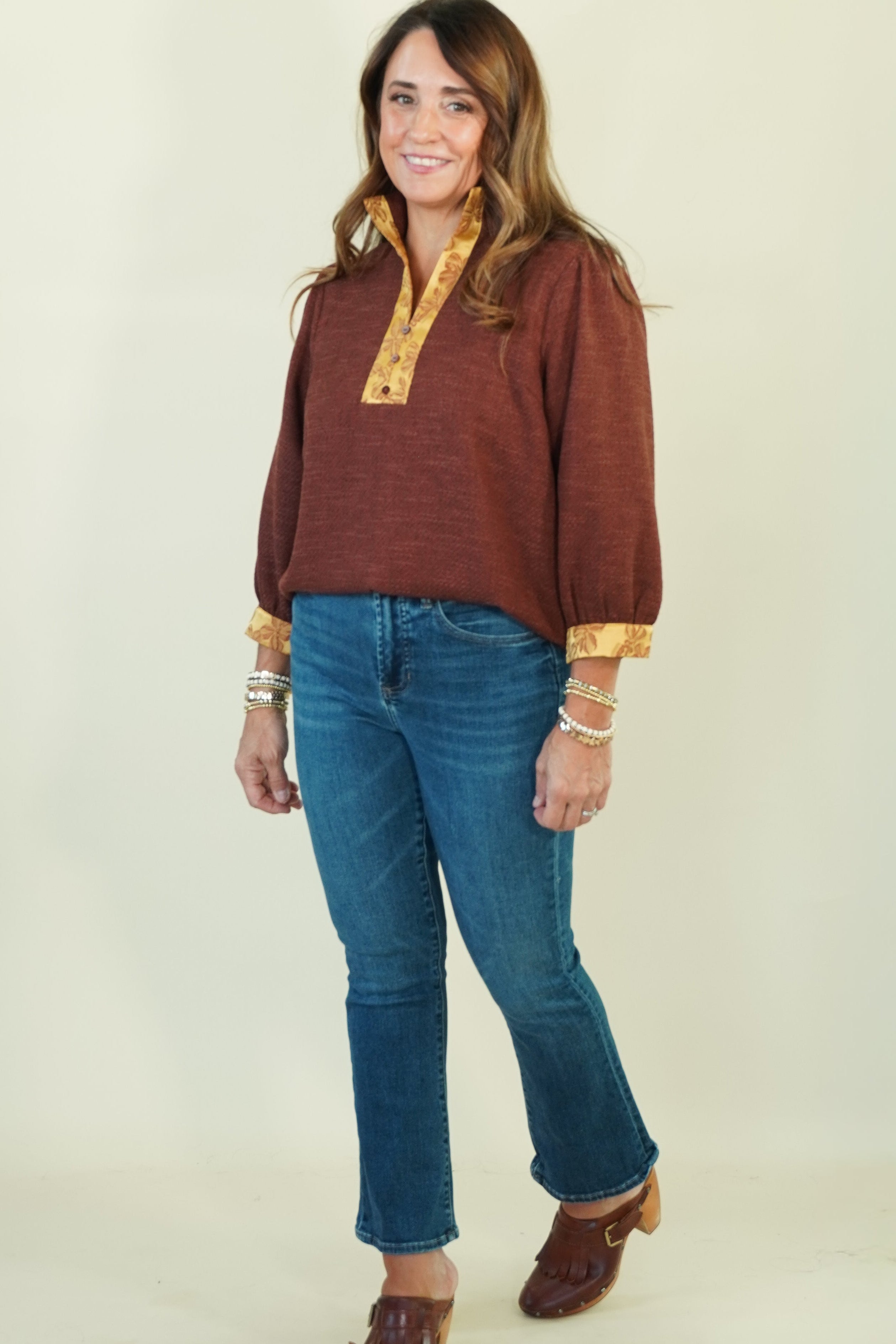 A woman modeling a rust-brown long-sleeve top with gold patterned trim on the collar and cuffs, paired with blue jeans and brown shoes, against a plain cream background.