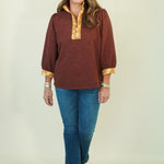 A woman modeling a rust-brown long-sleeve top with gold trim on the collar and cuffs, paired with blue jeans and brown shoes, against a plain cream background.
