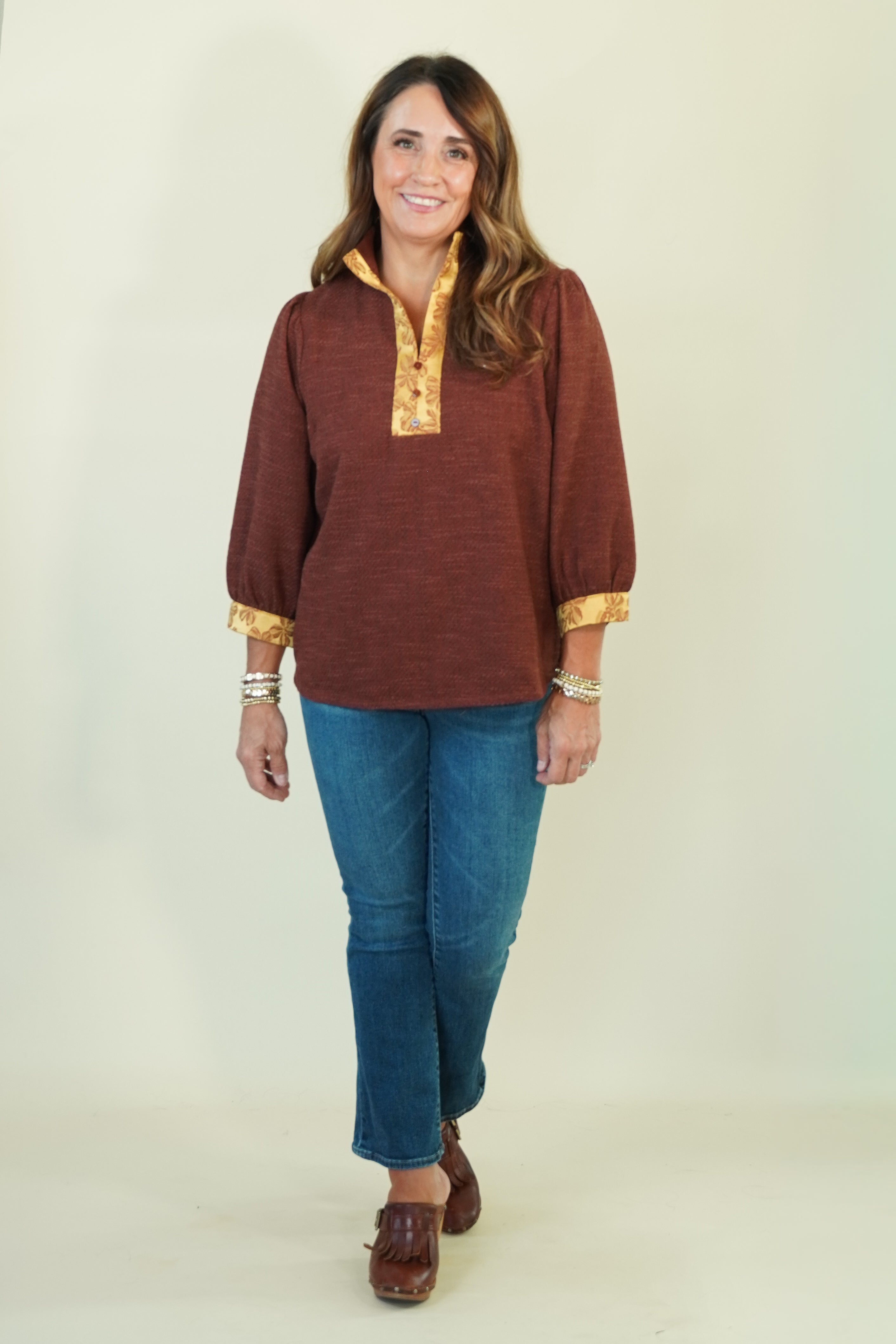 A woman modeling a rust-brown long-sleeve top with gold trim on the collar and cuffs, paired with blue jeans and brown shoes, against a plain cream background.