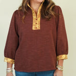 A woman modeling a rust-brown long-sleeve top with gold trim on the collar and cuffs, paired with blue jeans and brown shoes, against a plain cream background.