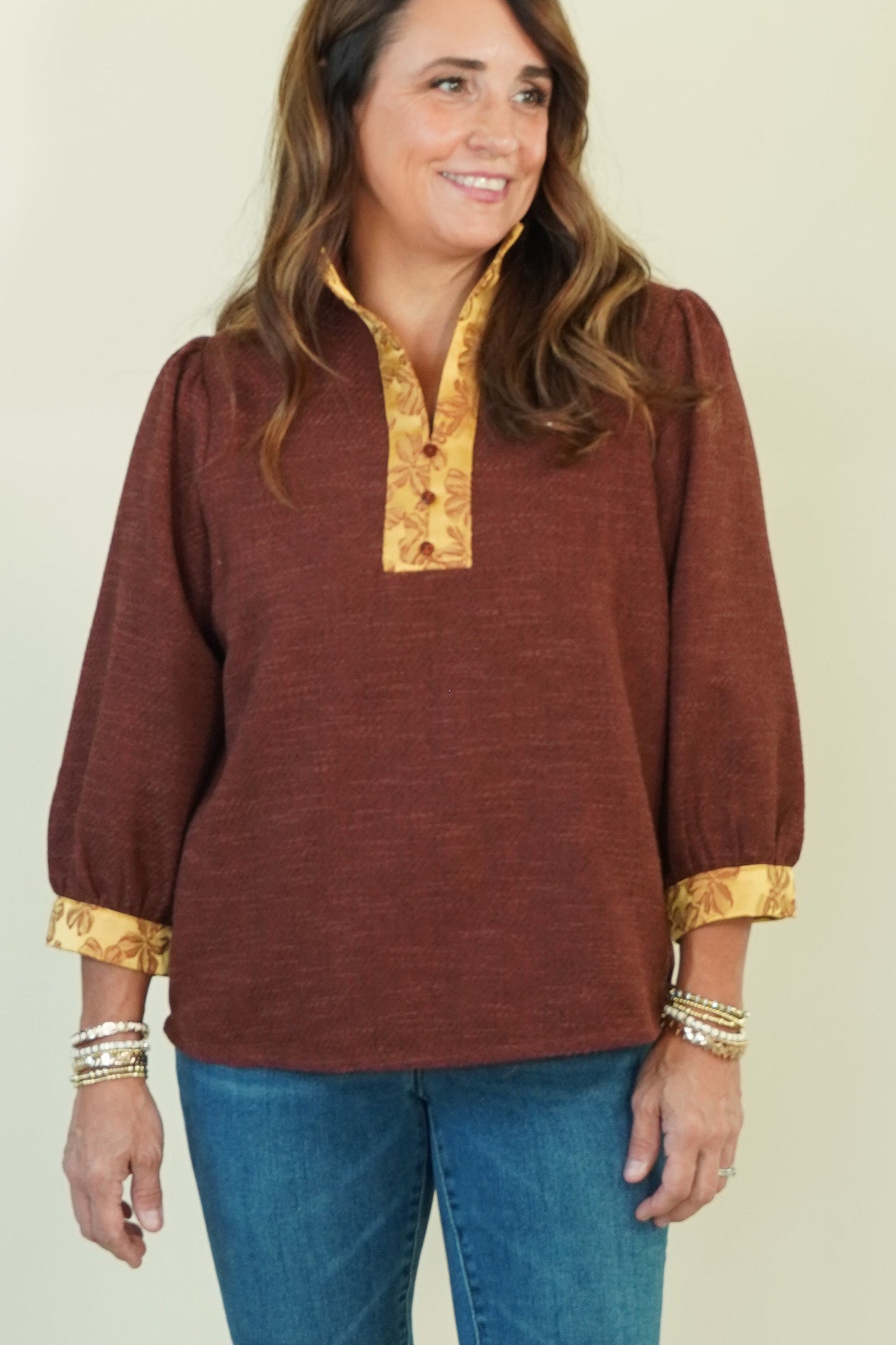 A woman modeling a rust-brown long-sleeve top with gold trim on the collar and cuffs, paired with blue jeans and brown shoes, against a plain cream background.