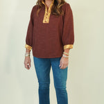 A woman modeling a rust-brown long-sleeve top with gold patterned trim on the collar and cuffs, paired with blue jeans and brown shoes, against a light cream background.