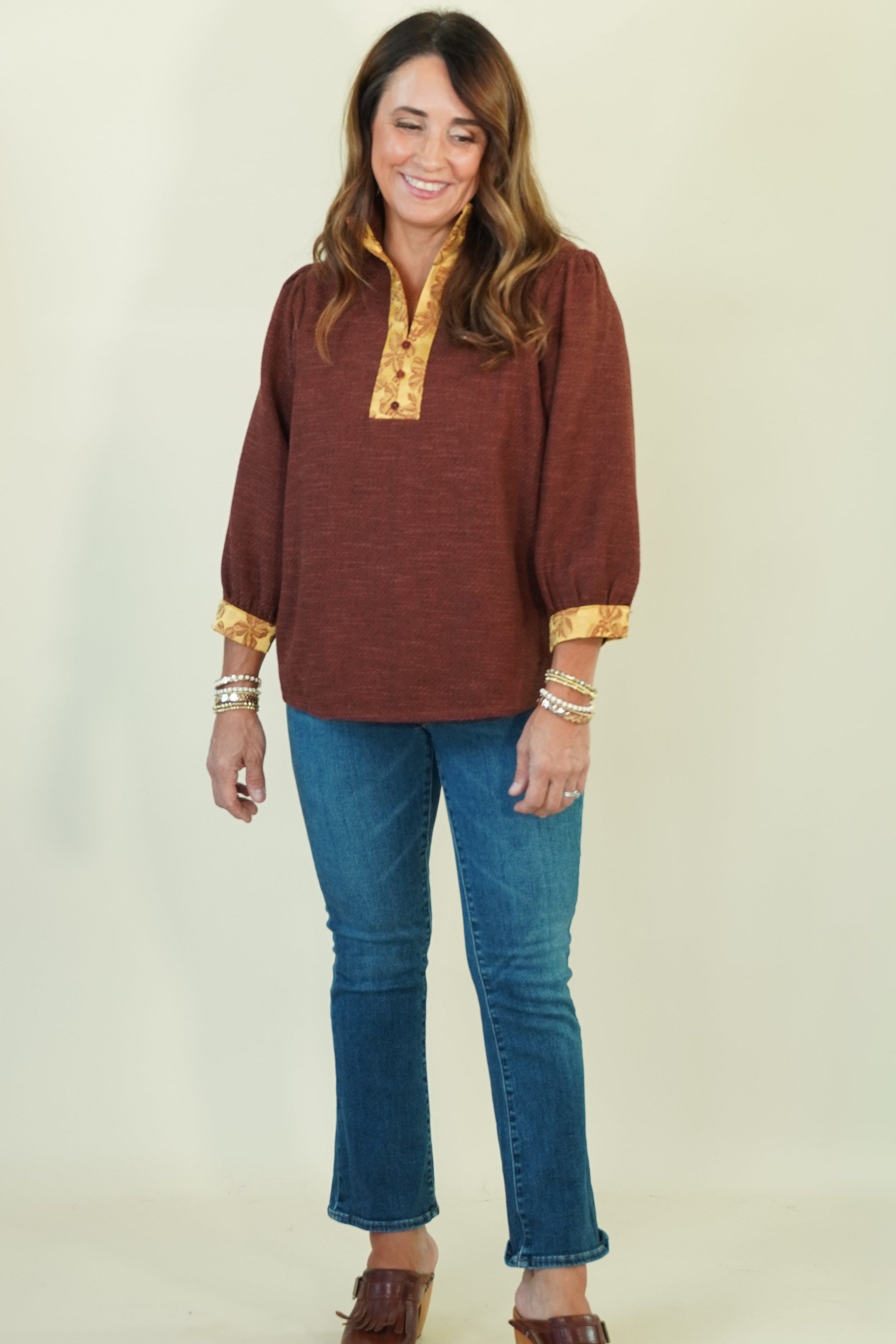 A woman modeling a rust-brown long-sleeve top with gold patterned trim on the collar and cuffs, paired with blue jeans and brown shoes, against a light cream background.