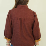 A woman viewed from the back wearing a rust-brown long-sleeve blouse with gold-trimmed cuffs, blue jeans, and brown heels, standing against a plain cream backdrop.