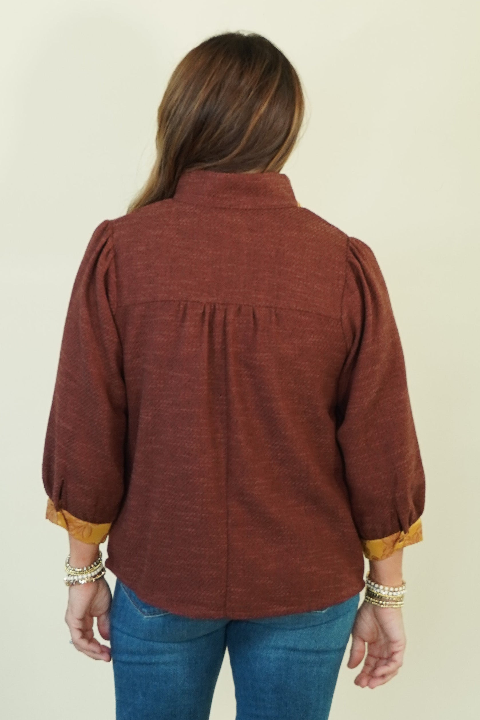 A woman viewed from the back wearing a rust-brown long-sleeve blouse with gold-trimmed cuffs, blue jeans, and brown heels, standing against a plain cream backdrop.