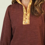 A woman modeling a rust-brown long-sleeve top with gold trim on the collar and cuffs, paired with blue jeans and brown shoes, against a plain cream background.