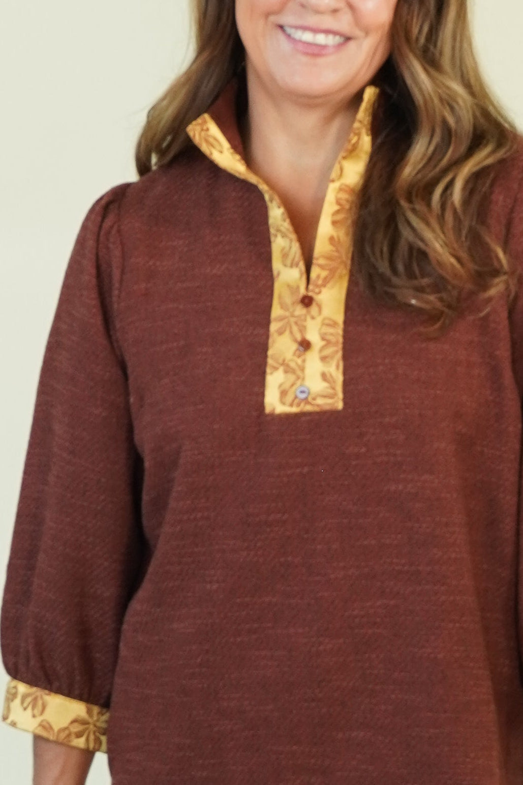A woman modeling a rust-brown long-sleeve top with gold trim on the collar and cuffs, paired with blue jeans and brown shoes, against a plain cream background.