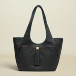 A black leather tote bag with a tassel detail and subtle contrasting stitching, photographed against a solid light beige background.
