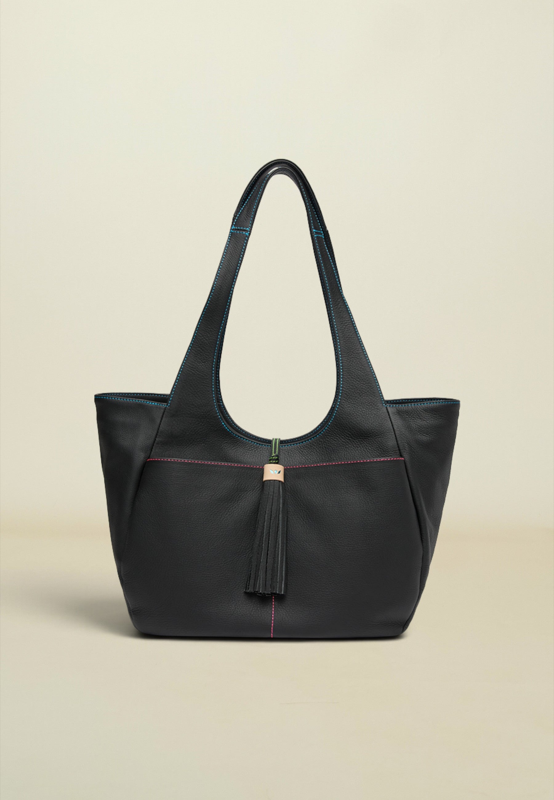 A black leather tote bag with a tassel detail and subtle contrasting stitching, photographed against a solid light beige background.