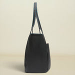A black leather tote bag with contrasting blue stitching on the handles, shown from a side profile view against a warm beige background.