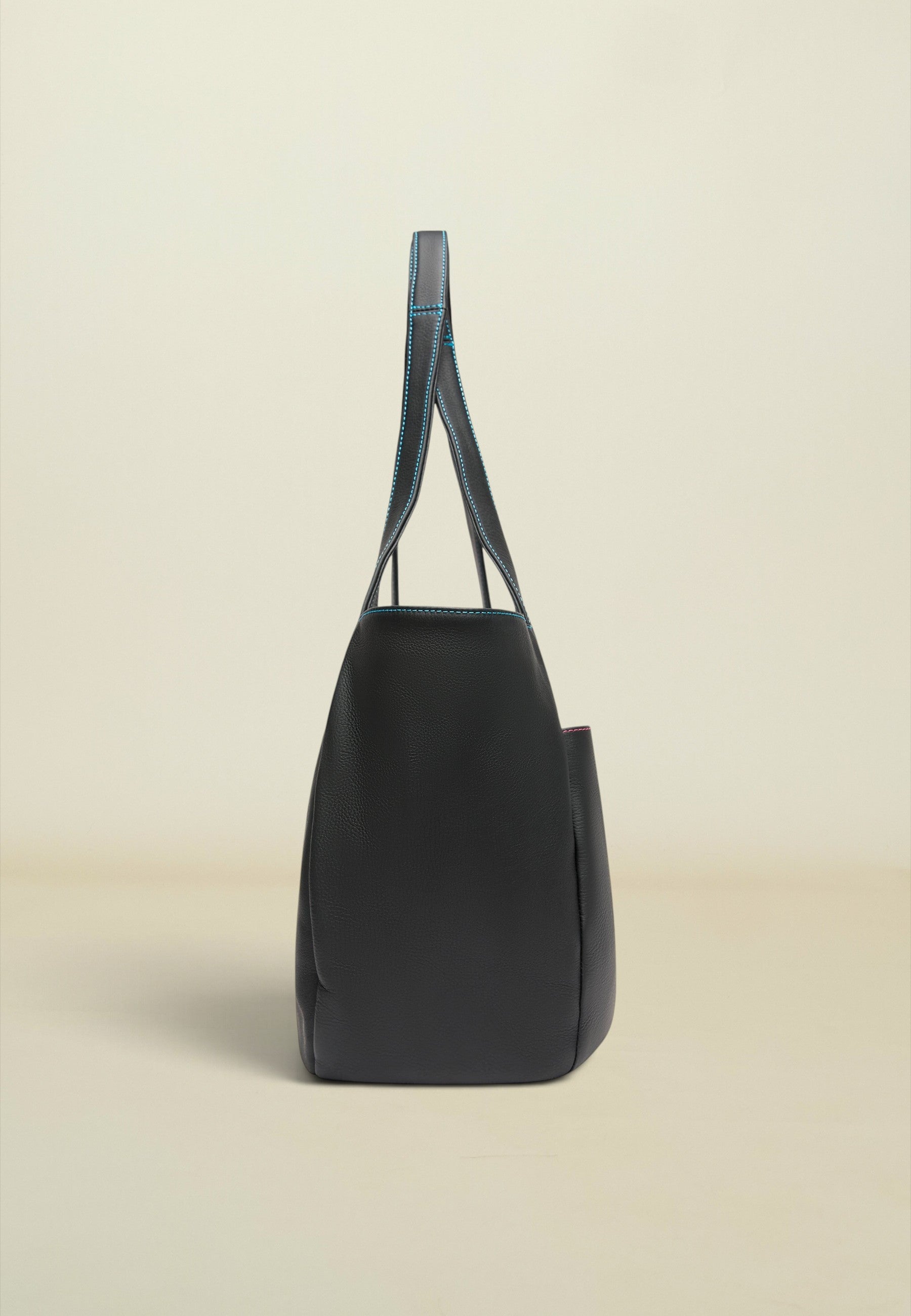 A black leather tote bag with contrasting blue stitching on the handles, shown from a side profile view against a warm beige background.