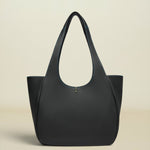 A structured black leather tote bag with a distinctive curved top handle, photographed against a solid, warm off-white background.