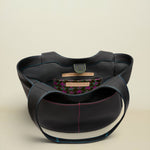 A black leather shoulder bag with a structured, rounded silhouette, featuring contrasting blue and pink stitching, a gold-tone metal ring closure, and a patterned interior label.