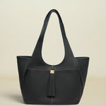 A structured black leather tote bag with double shoulder straps, a front tassel detail, and subtle pink contrast stitching, set against a solid light beige background.