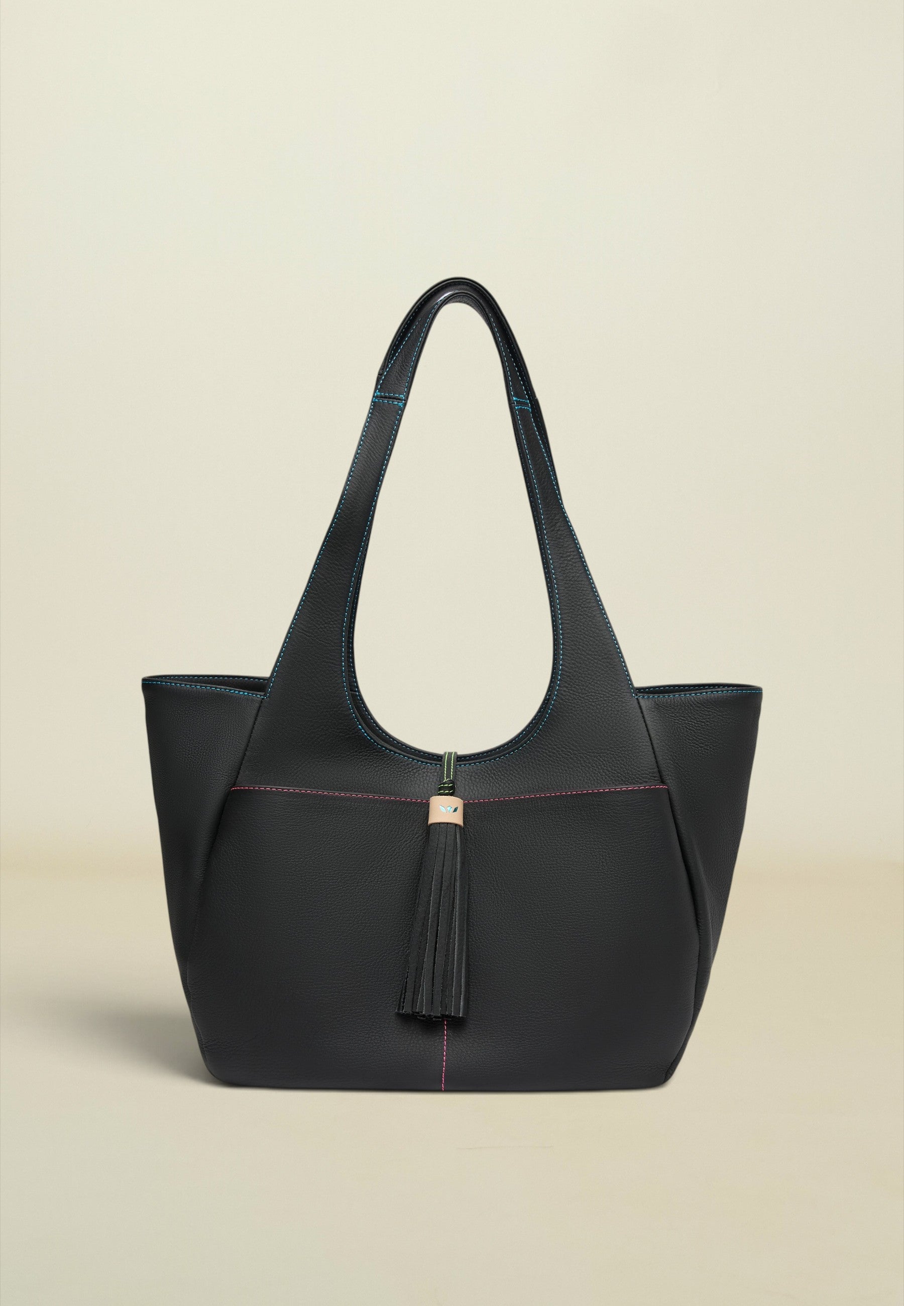 A structured black leather tote bag with double shoulder straps, a front tassel detail, and subtle pink contrast stitching, set against a solid light beige background.