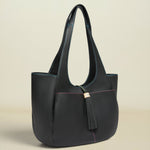 A structured black leather tote bag with long shoulder straps, a tassel detail, and subtle contrasting stitching, photographed against a solid light beige background.