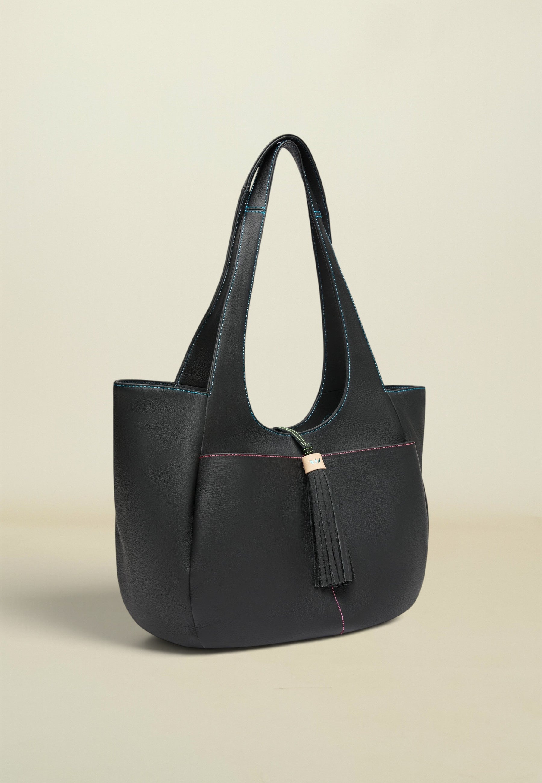 A structured black leather tote bag with long shoulder straps, a tassel detail, and subtle contrasting stitching, photographed against a solid light beige background.