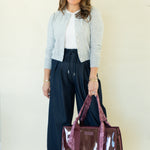 A woman stands against a plain light beige studio background wearing a light gray cardigan, white top, dark navy wide-leg pants, and white sneakers, while holding a large glossy burgundy tote bag with pink handles.