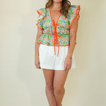 A woman modeling a vibrant tropical print ruffle top with orange trim and white shorts against a solid beige studio background.