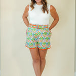 A woman modeling a white sleeveless top and colorful patterned shorts with a white and metallic slide sandal, standing against a plain beige studio background.