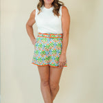 A woman modeling a pair of colorful, patterned shorts with a white sleeveless top and white slide sandals, standing against a plain beige studio background.