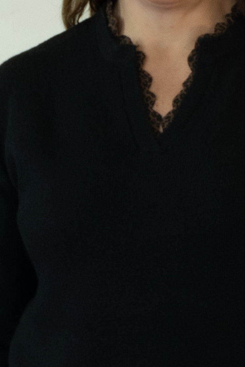 A woman modeling a black long-sleeve knit sweater with a split neckline, paired with white pants and brown shoes, standing against a plain off-white studio backdrop.