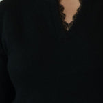 A woman modeling a black long-sleeve knit sweater with a split neckline, paired with white pants and brown shoes, standing against a plain off-white studio backdrop.