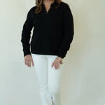 A woman modeling a black long-sleeve knit top with a split neckline, paired with white pants and brown shoes, standing against a plain off-white studio backdrop.
